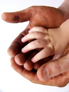 Image of large hands darkened with grease holding a single small clean hand. White background. Vertical orientation. ** Note: Slight blurriness, best at smaller sizes