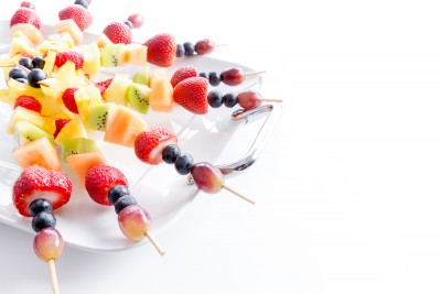 Serving of colorful healthy tropical fresh fruit kebabs with an assortment of exotic fruit on a white platter for a tasty vegan or vegetarian buffet over white with copy space