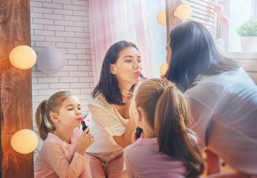 Funny family at home. Mother and her child girl are doing your makeup and having fun near mirror. Mom and daughter wear lipstick in kids room.